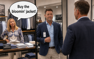 Man trying on a £249 jacket in a clothes shop mirror while his wife says, “Buy the bloomin’ jacket!”